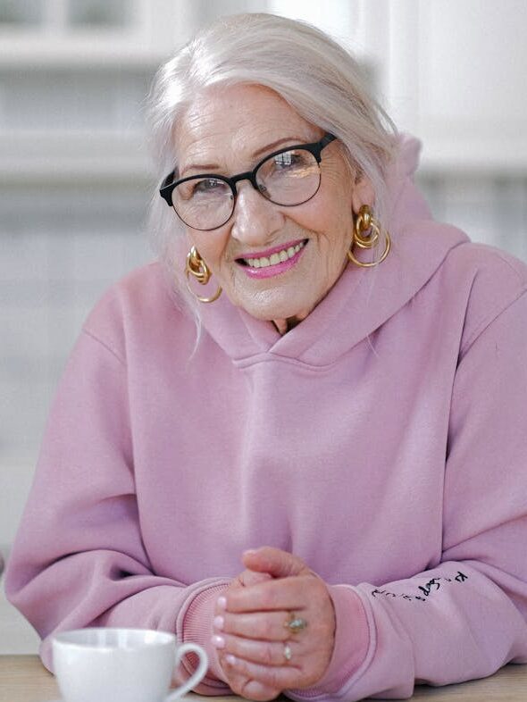 Smiling aged woman in hoodie sitting at table in light apartment